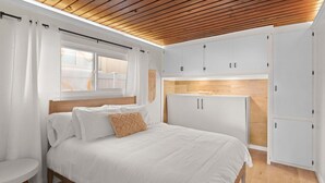 A bedroom with a wooden ceiling, a bed with white bedding, a small table, and white cabinets.