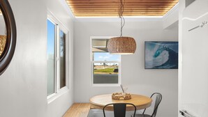 Ein Essbereich mit einem runden Holztisch, zwei schwarzen Stühlen, einer gewebten Pendelleuchte und einem Bilderrahmen mit einer Wellenansicht an der Wand.