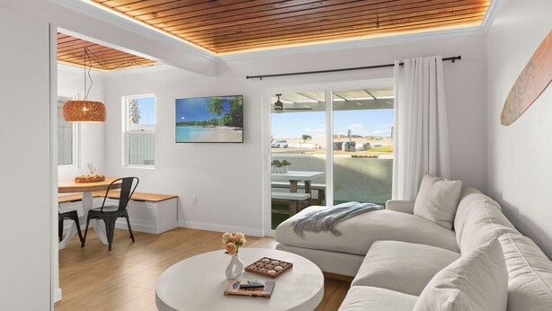 Ein modernes Wohnzimmer mit Holzboden, einem weißen Sofa, einem runden Couchtisch mit Vase und Büchern, einem Essbereich mit Holztisch und Stühlen sowie einem großen Fenster mit Blick auf den Strand.
