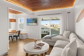 A modern living room with a wooden ceiling, a white sofa, a round coffee table with a vase and books, a dining area with a wooden table and chairs, and a large window with a view of the beach. - New! 1 bd/1 ba Beach Front Bungalow (San Diego)