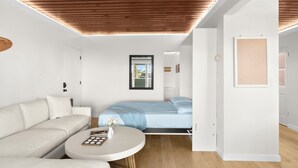 Ein modernes, minimalistisches Schlafzimmer mit Holzbalkendecke, einem Bett mit blauer Bettdecke, einem weißen Sofa und einem kleinen runden Tisch mit Blumen.'