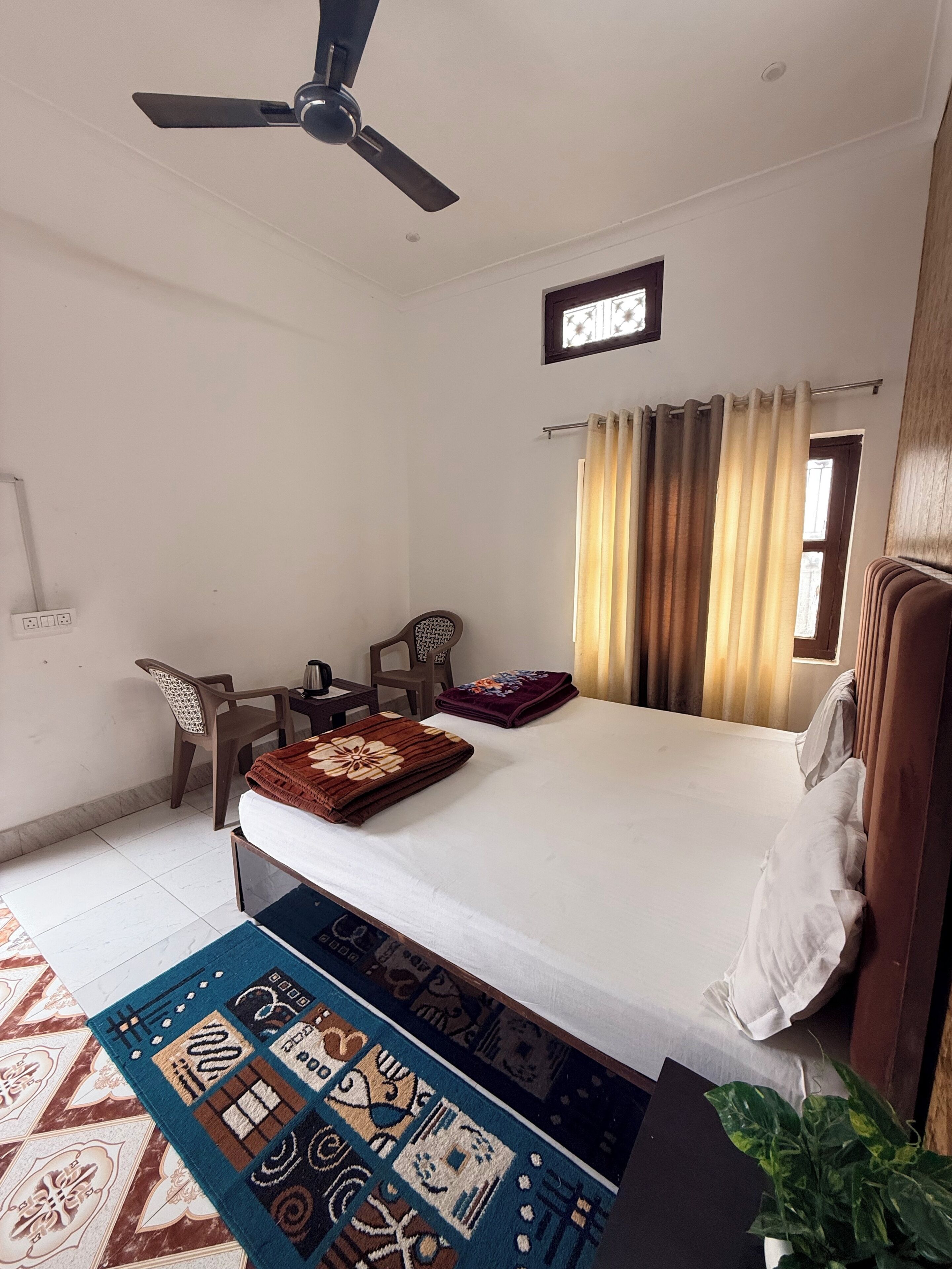 Habitación de hotel con cama, ventilador de techo, escritorio, dos sillas, ventana con cortinas y una planta en maceta.