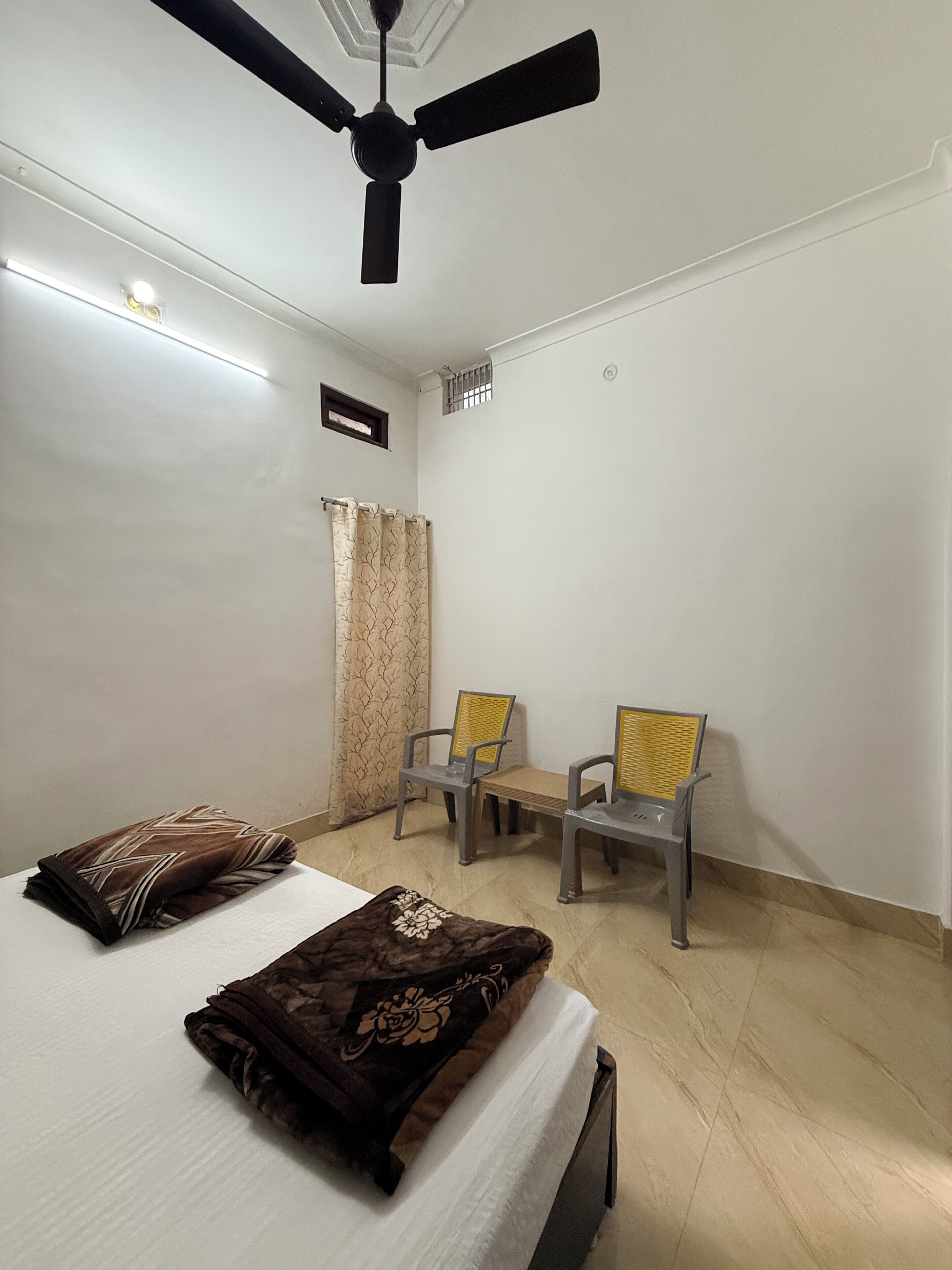 A room with a bed, two chairs, a ceiling fan, and a window with curtains.