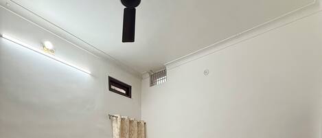 A room with a bed, two chairs, a ceiling fan, and a window with curtains.
