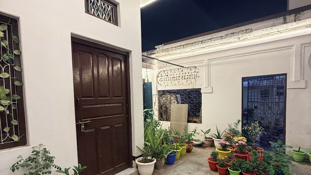A courtyard with a variety of potted plants, a brown door, and a white wall with decorative elements. - Saryu Sankul Homestay (Ayodhya)