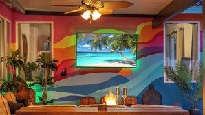 A room with a TV displaying a beach scene, a wooden table with two glasses, and a ceiling fan.