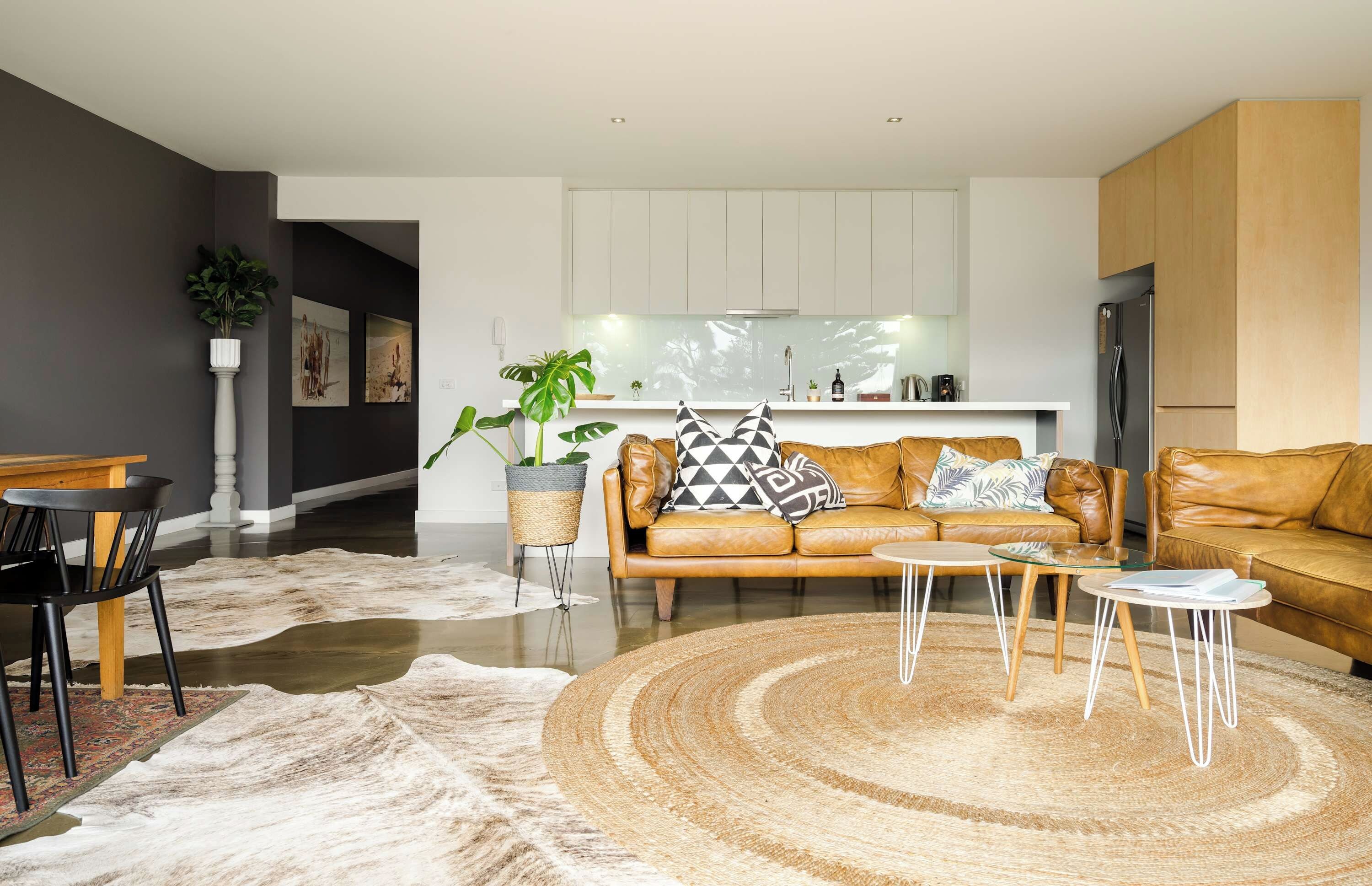 Ein modernes Wohnzimmer mit einer Ledersofa, einem Couchtisch und einem Teppich. Im Hintergrund ist eine Küche mit weißen Schränken und einem Kühlschrank zu sehen.
