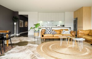 A modern living room with a leather sofa, a coffee table, and a rug. There is a kitchen in the background with white cabinets and a refrigerator.