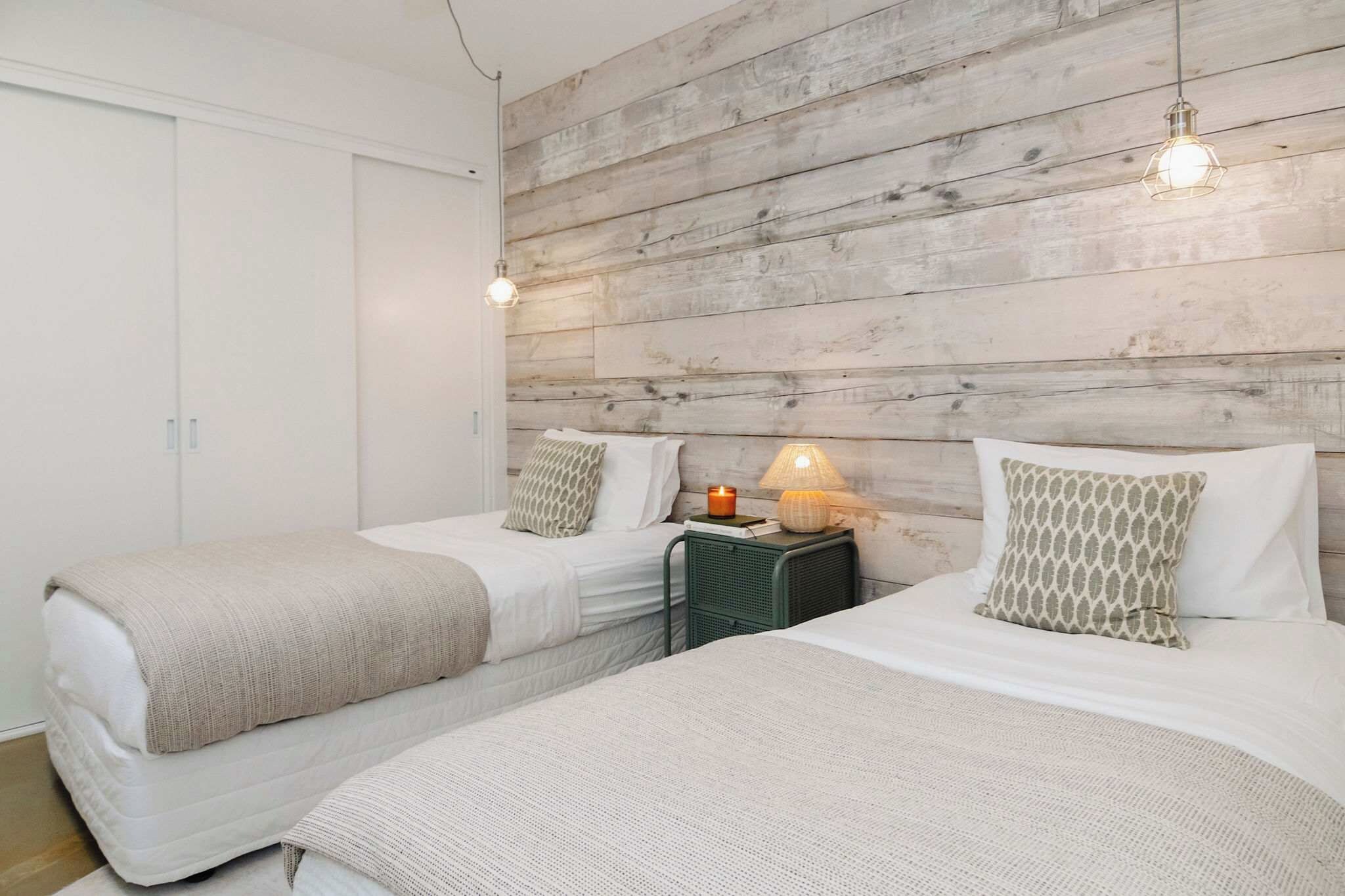 A bedroom with two beds, a wooden wall, a nightstand with a lamp, and a white wardrobe.