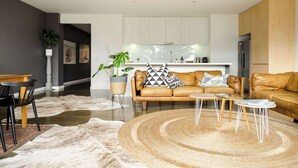A modern living room with a leather sofa, a coffee table, and a rug. There is a kitchen in the background with white cabinets and a refrigerator. - Aquabelle One on Rye Beach (Rye)