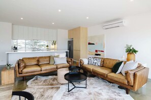 A modern living room with two brown leather sofas, a round coffee table, and a fur rug. There is a kitchen area with white cabinets and a refrigerator.