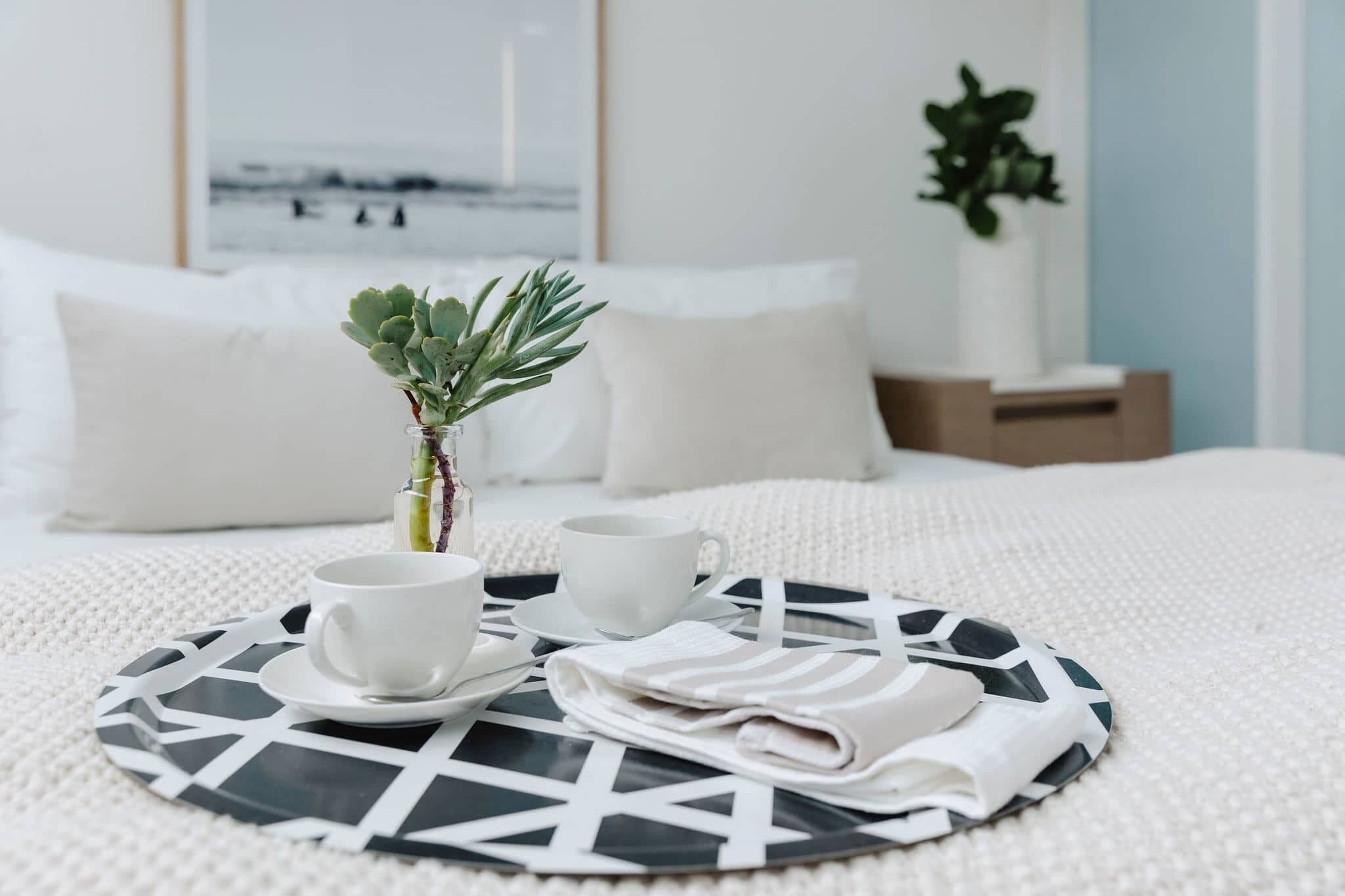 A neatly made bed with a white comforter, a small potted plant, two white cups, and a folded towel on a black and white patterned tray.