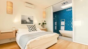A modern bedroom with a large bed, bedside tables, a wall-mounted air conditioner, a framed picture, a potted plant, and a bathroom with a blue tiled wall and a white bathtub.