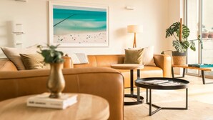 A modern living room with a beige sofa, a round coffee table, a side table, and a framed beach painting on the wall.