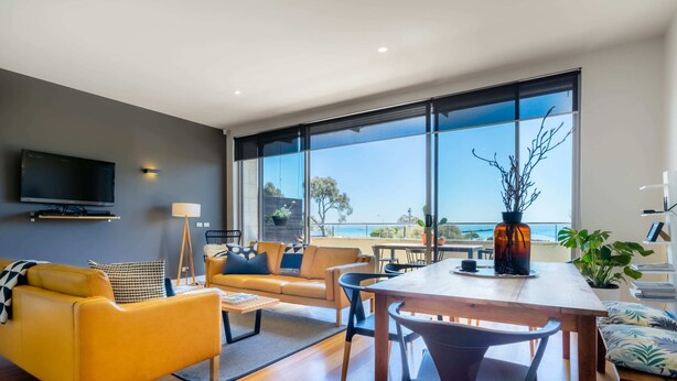 A modern living room with a sofa, chairs, a coffee table, and a dining table. There is a TV mounted on the wall and a large window offering a view of the outdoors. - Aquabelle Four on Rye Beach (Rye)