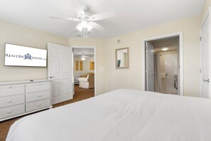 Une chambre à coucher avec un lit, une commode, un ventilateur de plafond, une porte et une salle de bain visible à travers une porte ouverte.