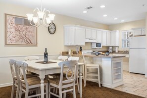 Une cuisine avec des meubles blancs, une table à manger avec des chaises, un lustre et une horloge au mur.