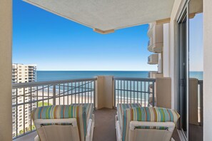 Een balkon met twee gestreepte stoelen, een helder uitzicht op het strand en een hoog gebouw op de achtergrond.