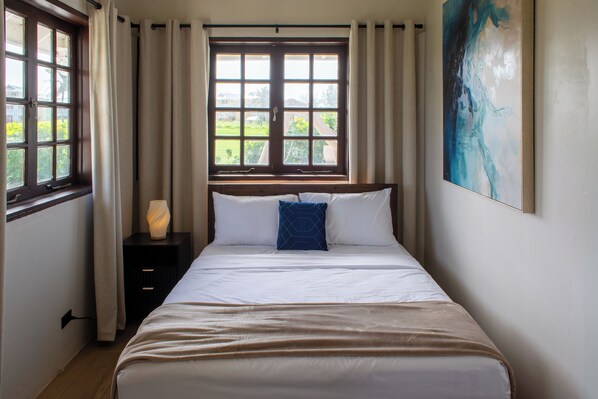 Habitación de hotel con un ventanal grande, cama con ropa blanca, almohada azul, mesita de noche con lámpara y un cuadro en la pared.