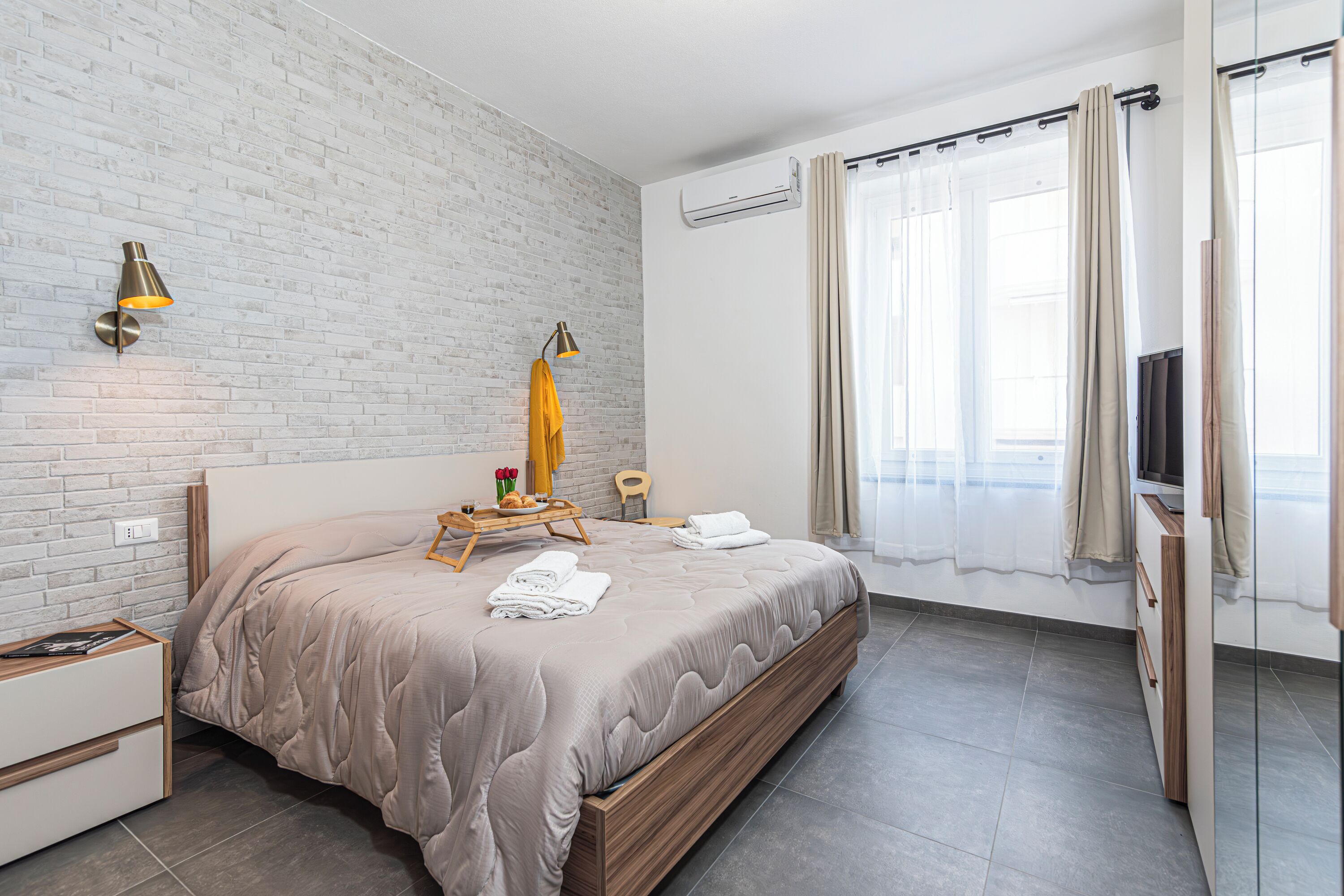 A modern hotel room with a brick wall, a large bed with a wooden frame, a bedside table, a small wooden chair, and a window with curtains.