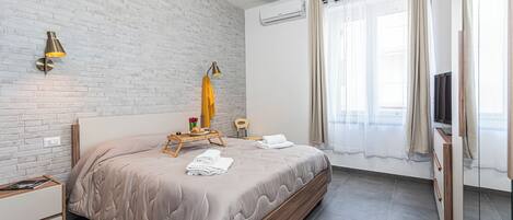 A modern hotel room with a brick wall, a large bed with a wooden frame, a bedside table, a small wooden chair, and a window with curtains.