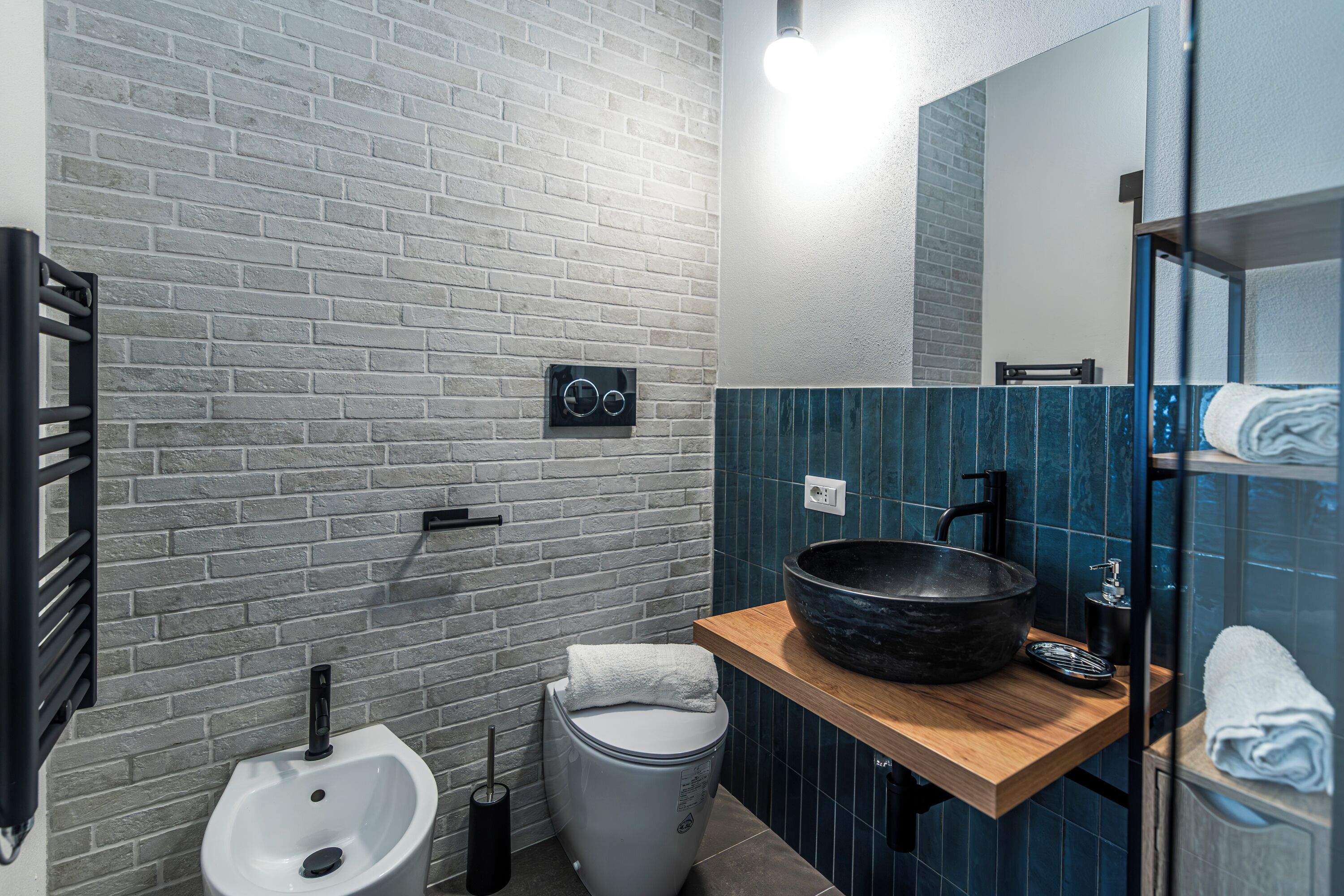 A modern bathroom with a white brick wall, a wooden vanity, a large black basin, a toilet, and a towel rack.