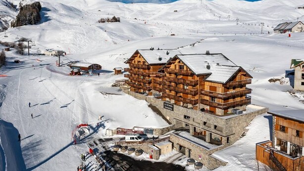 Een houten lodge met meerdere balkons, gelegen op een besneeuwde berghelling met skipistes op de achtergrond.
