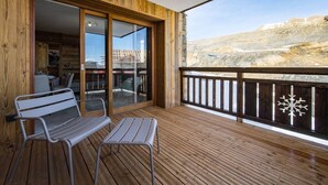 Een houten terras met een bankje en een tafel, een glazen deur die leidt naar een binnenruimte, en uitzicht op een bergachtig landschap.