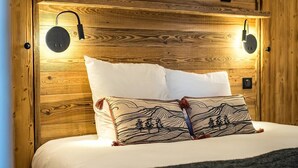 Een slaapkamer met een houten hoofdbord, twee bedlampjes, een bed met wit beddengoed en een sierkussen met een patroon.
