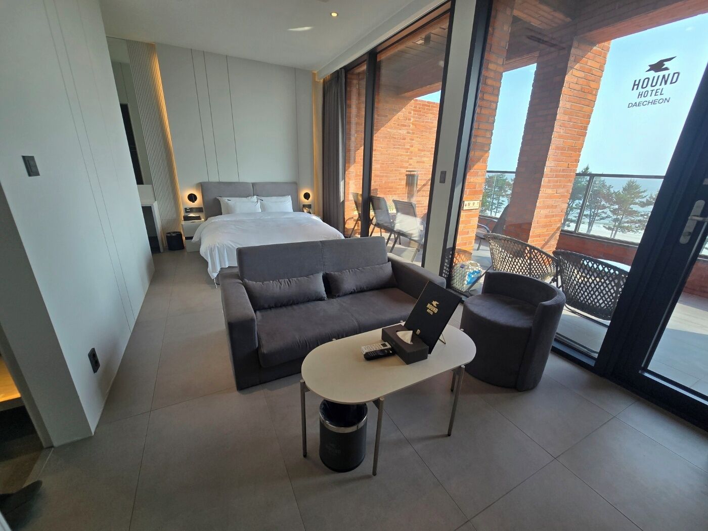 A modern hotel room with a large bed, a sofa, a small table, and a balcony with a view of the outside.
