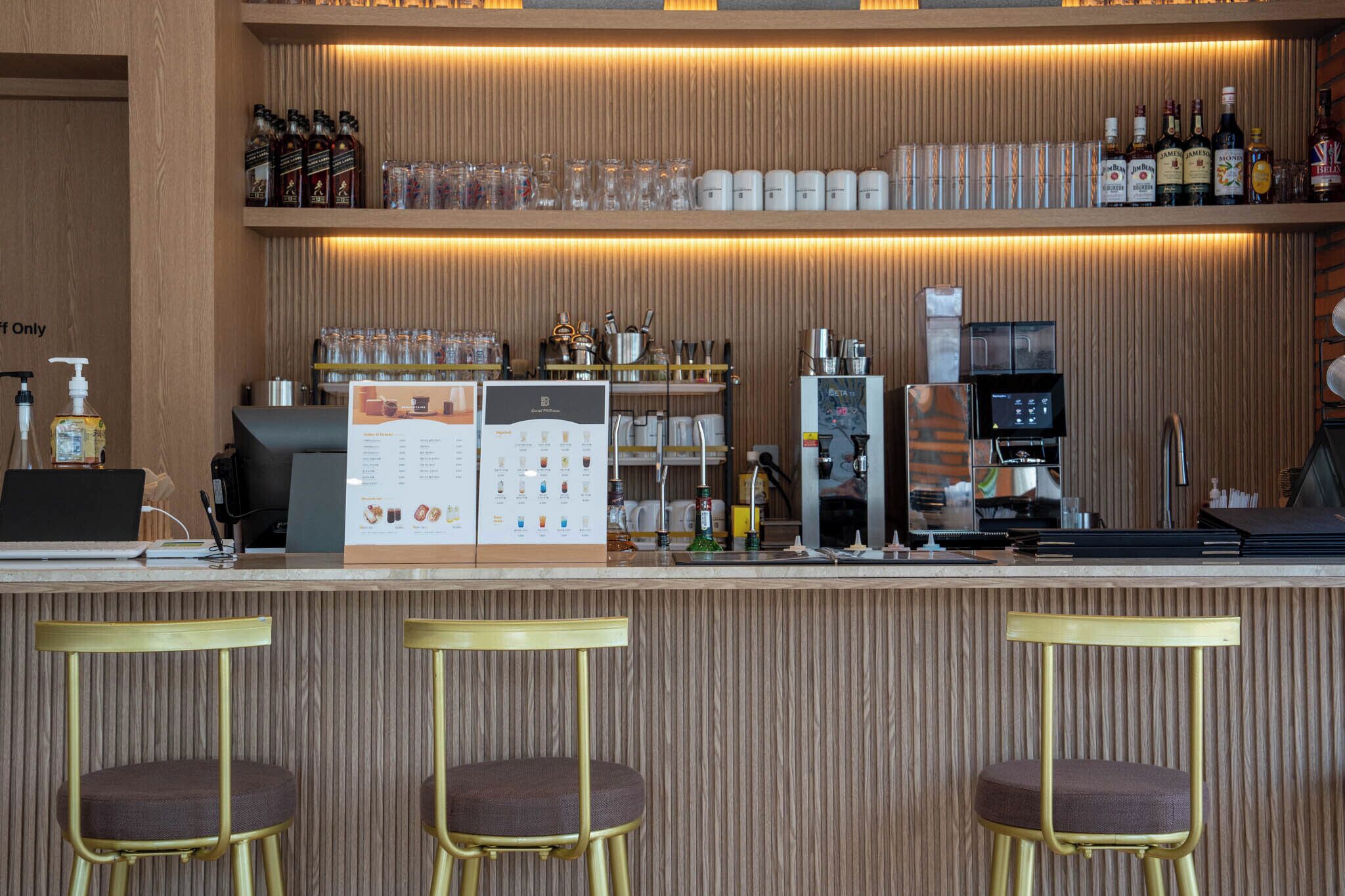 Una zona de bar con taburetes, una máquina de café y estantes con botellas y vasos.