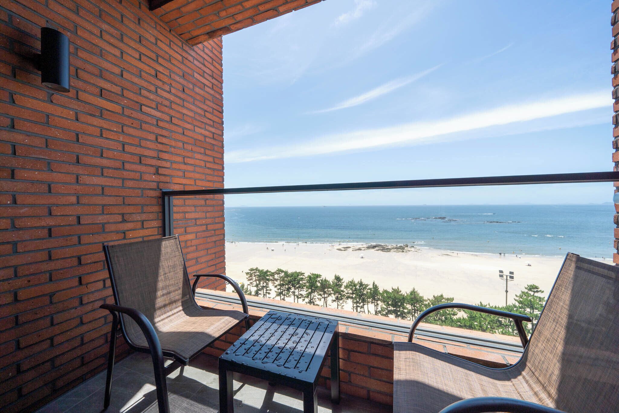 A balcony with a view of the beach, featuring a brick wall, two chairs, and a table.