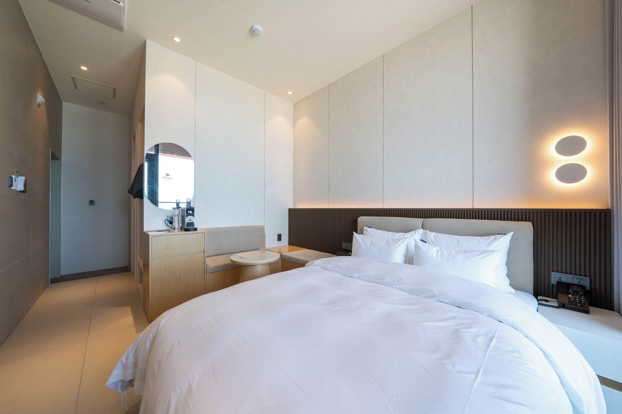 A modern hotel room with a large bed, bedside tables, a mirror, and a wall-mounted light fixture.