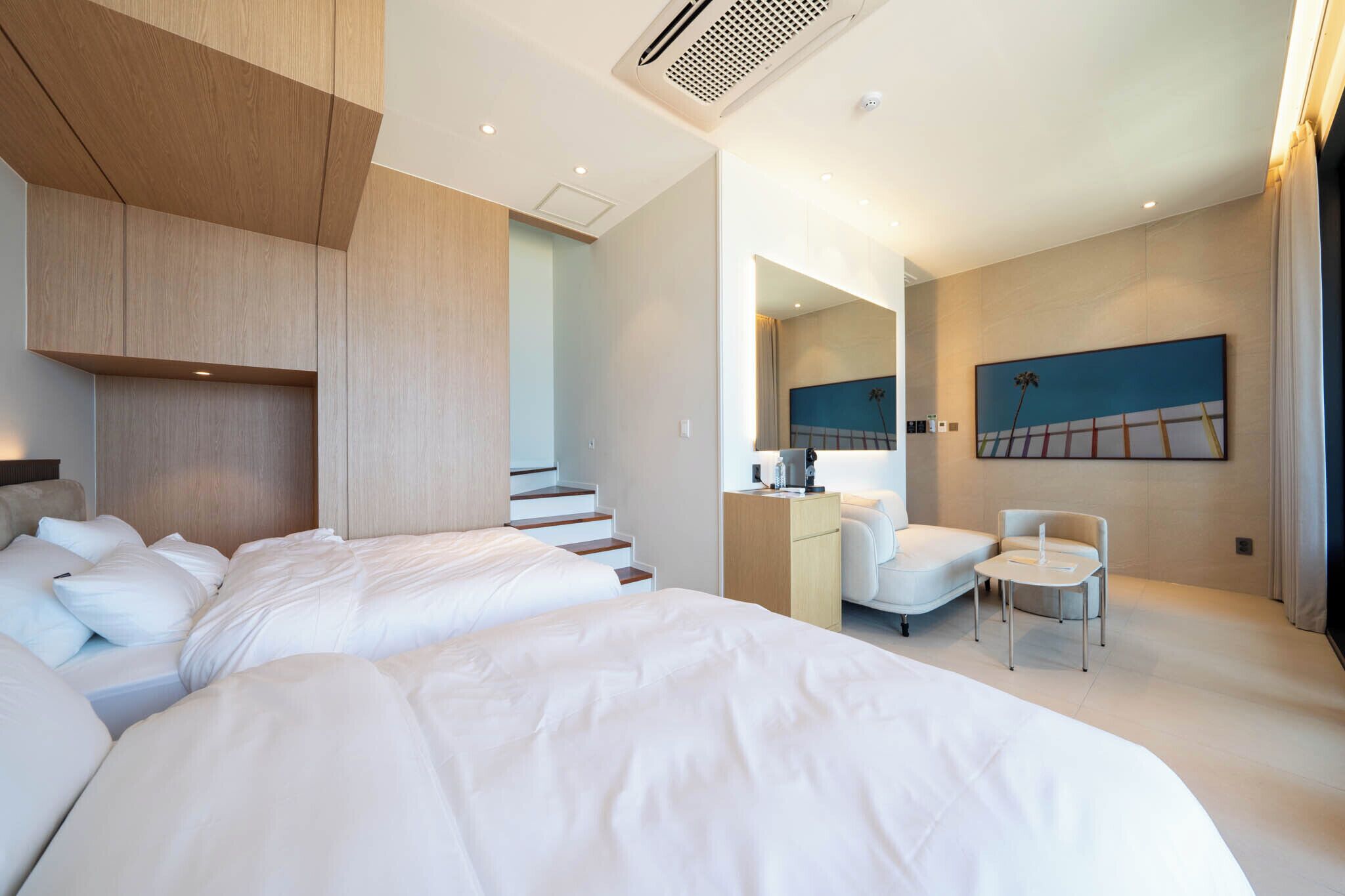 A modern hotel room with a large bed, a desk, a chair, and a staircase leading to an upper level.