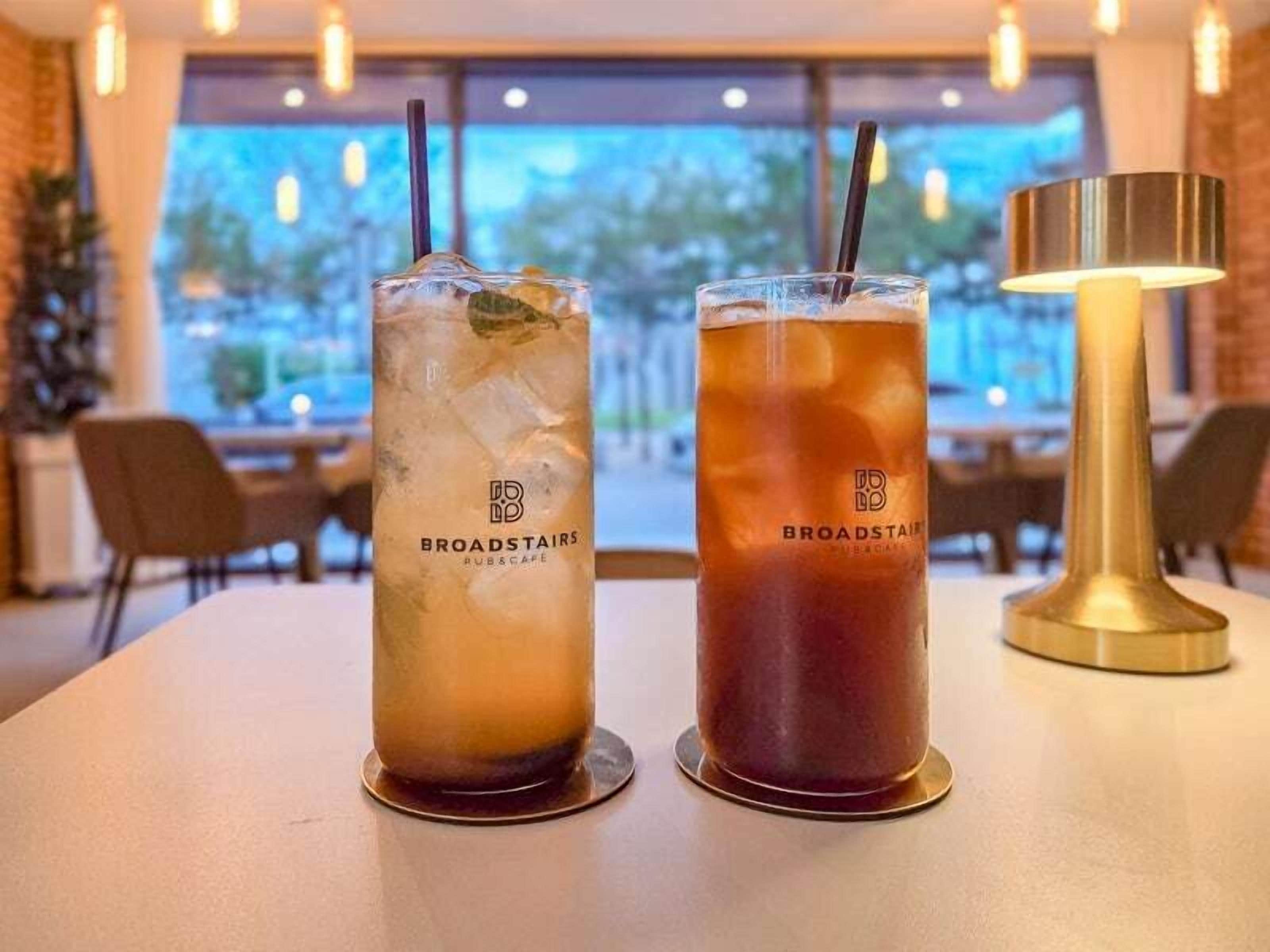 Two glasses of iced tea with a straw and the Broadstairs logo on a table in a restaurant with a view of the outdoors.