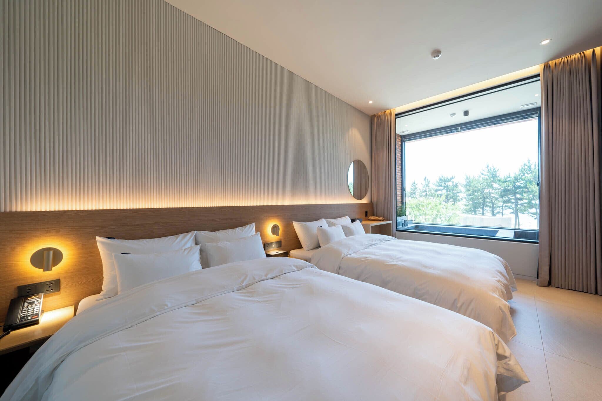 A modern hotel room with two beds, a large window with curtains, a round wall-mounted light, and a telephone on the nightstand.