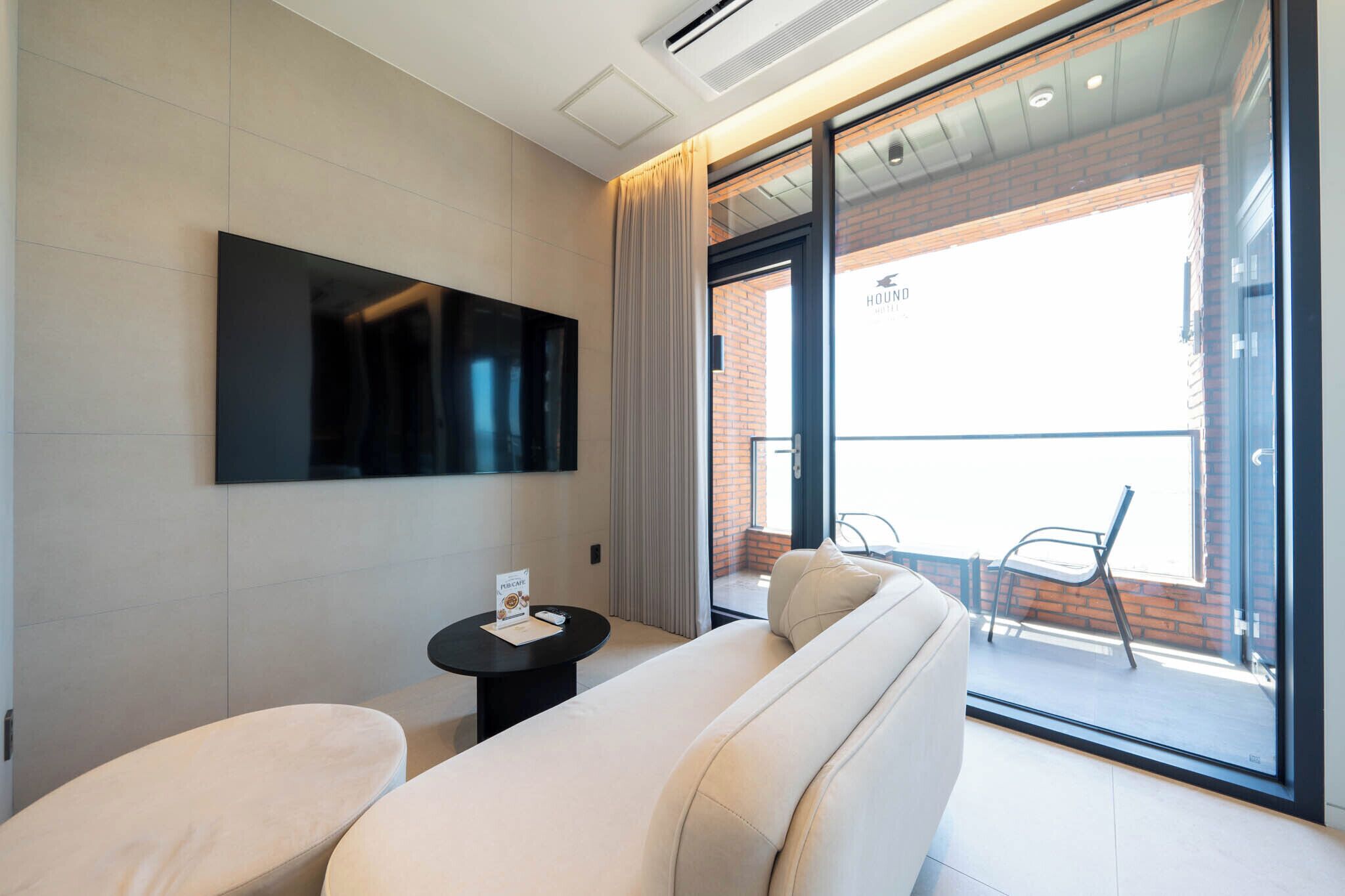 A modern hotel room with a large flat-screen TV mounted on the wall, a sofa, a small table with a menu, and a sliding glass door leading to an outdoor balcony with a chair.