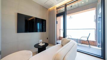 A modern hotel room with a large flat-screen TV mounted on the wall, a sofa, a small table with a menu, and a sliding glass door leading to an outdoor balcony with a chair.
