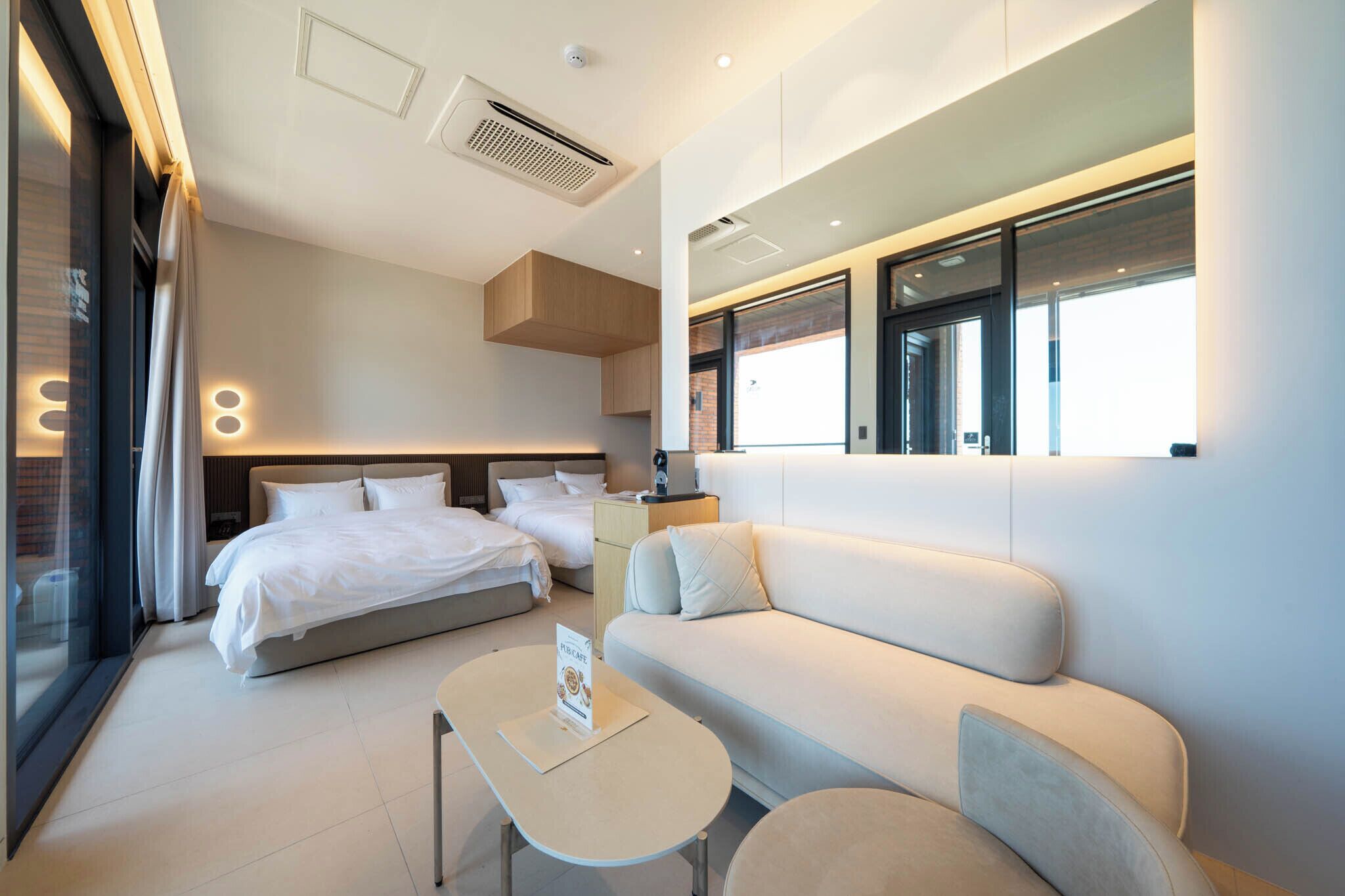 A modern hotel room with a large bed, a sofa, a small table, and a glass partition.