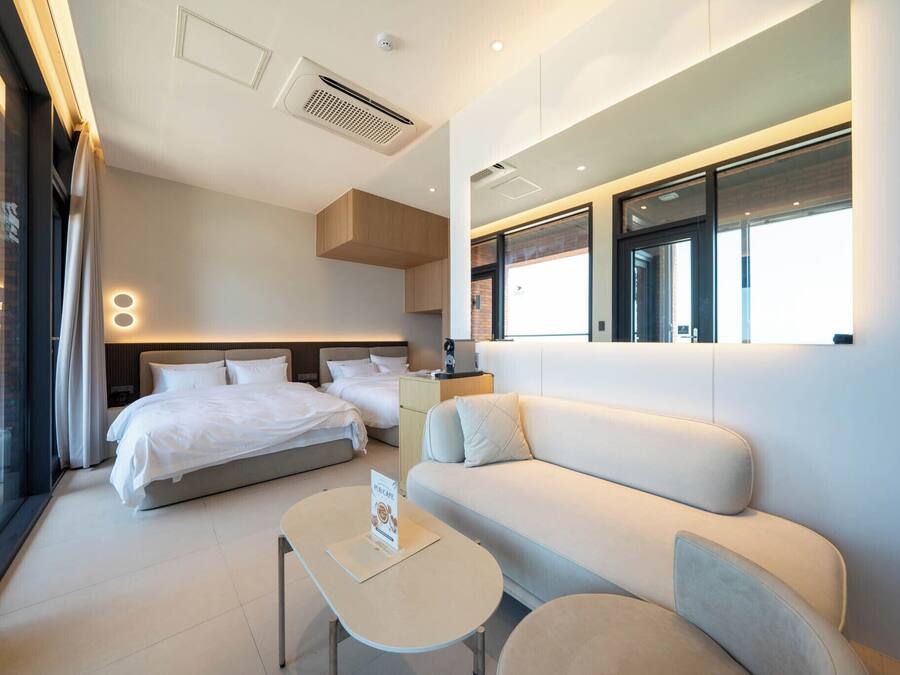 A modern hotel room with a large bed, a sofa, a small table, and a glass partition.