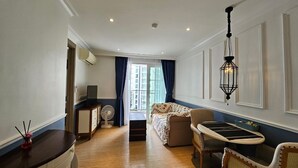 A well-lit room with a sofa, a dining table, and chairs. There is a fan on the floor and a window with curtains.