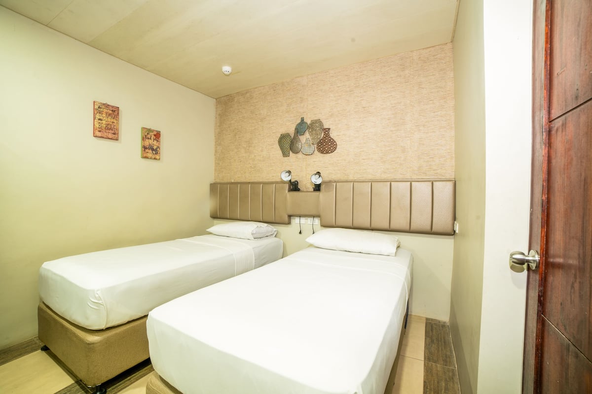 A hotel room with two single beds, a wooden headboard, and a wall-mounted light fixture.
