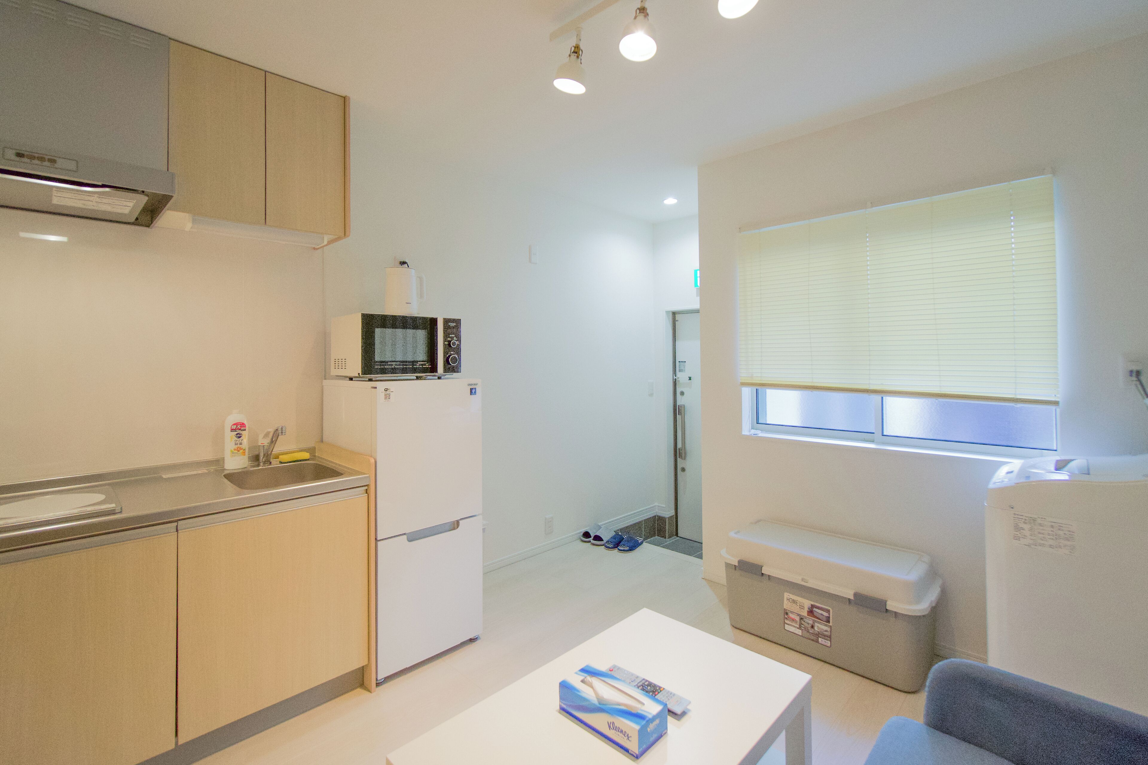 A compact kitchen with a microwave, sink, and cabinets. There's a small table with a box on it and a window with blinds.