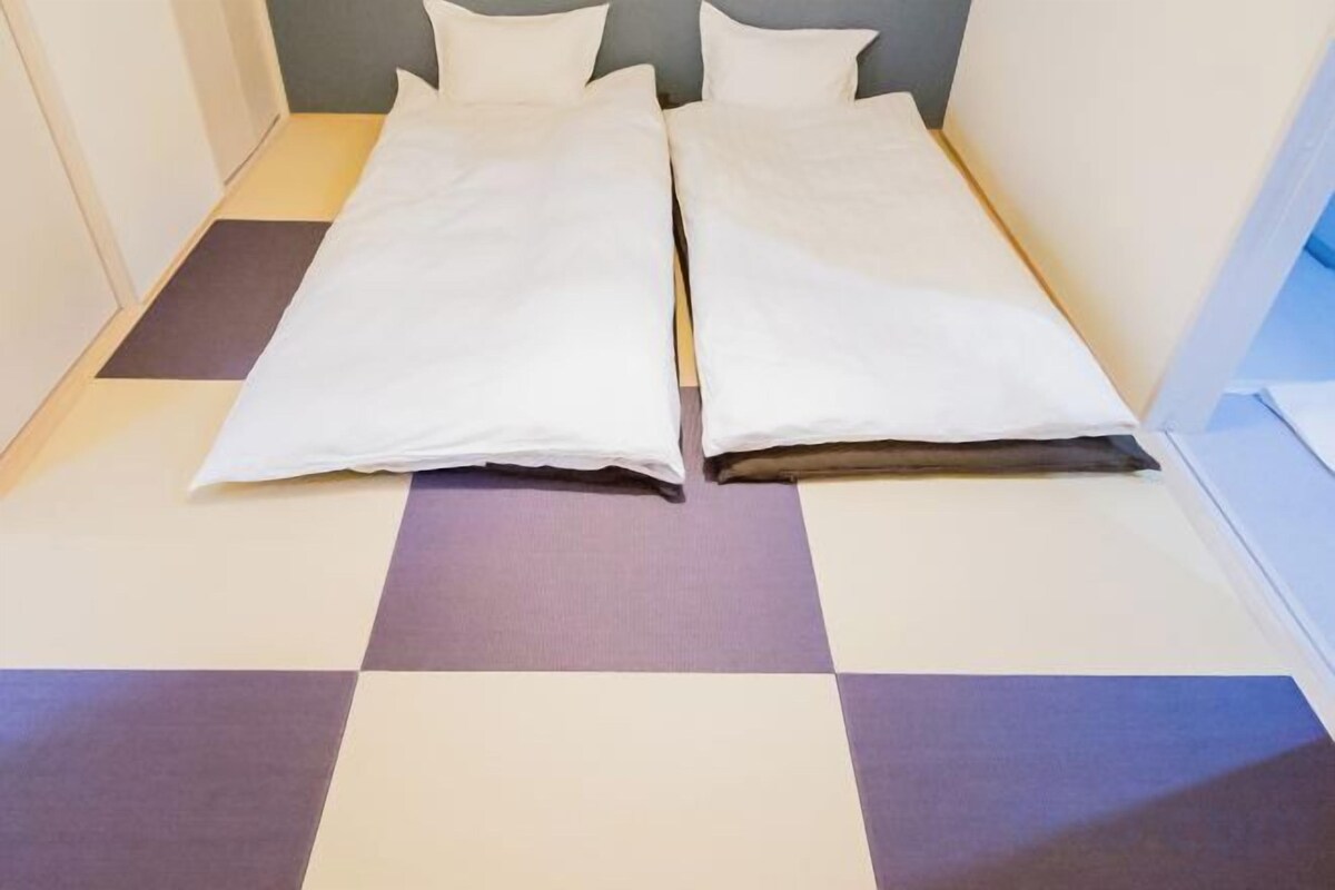 Una cama con sábanas blancas, piso de cuadros y cabecera sencilla.