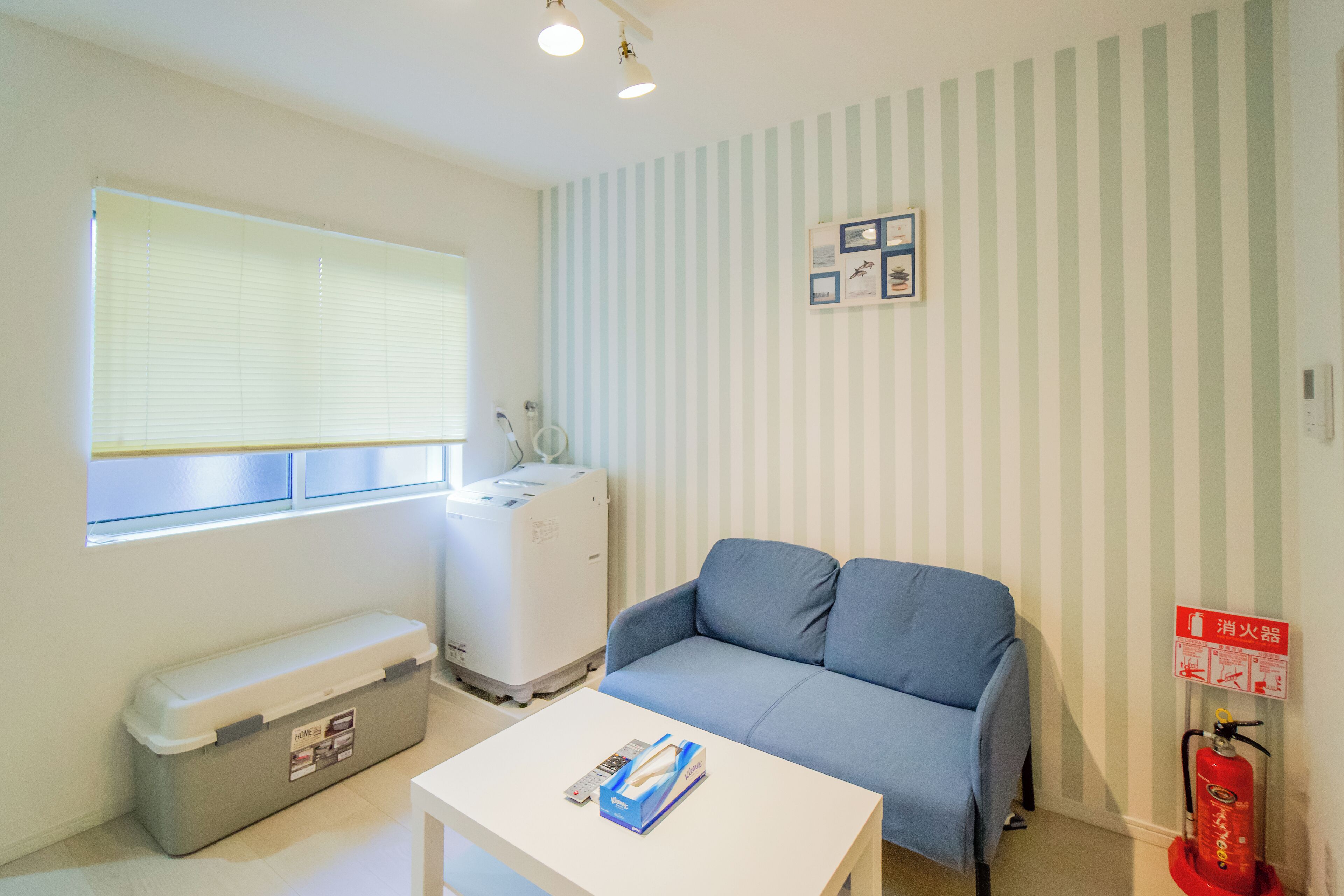 Une pièce avec un canapé bleu, une table blanche, un meuble de rangement, une fenêtre avec des stores, un appareil électroménager blanc et un extincteur.