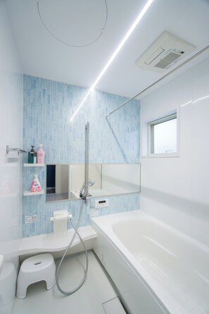 Un bagno moderno con una vasca da bagno bianca, una doccia e uno sgabello. Sono presenti mensole con articoli da toilette e una finestra sopra la vasca.