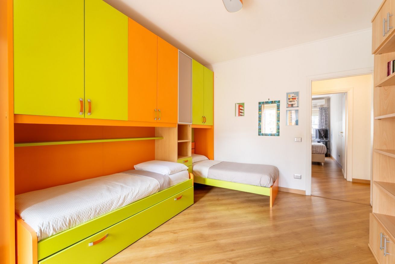 A room with two beds, bright orange and yellow cabinets, wooden flooring, and a visible doorway leading to another room.