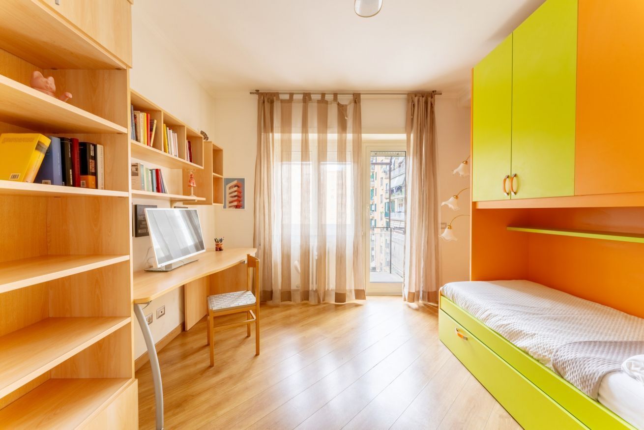 Een slaapkamer met een bed, een bureau, een stoel, een boekenkast en een raam met gordijnen.