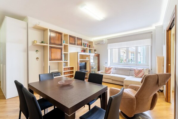 A modern living room with a dining table, chairs, a sofa, and a recliner. There is a built-in bookshelf and a television.