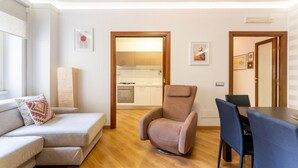Un salon comprenant un canapé, un fauteuil inclinable et une table à manger avec des chaises. On peut apercevoir la cuisine à travers une porte ouverte.